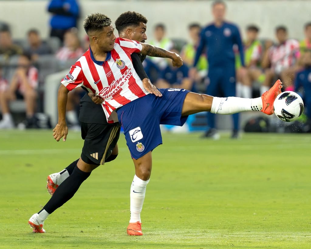 Efraín Álvarez, in Guadalajara's red and white striped jersey, battling an LAFC player for the ball as he clears it with a high kick.