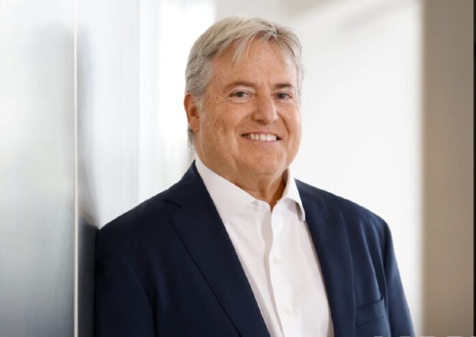Larry Berg, smiling, wearing a navy blazer and open-collar white shirt, standing indoors near a glass wall.