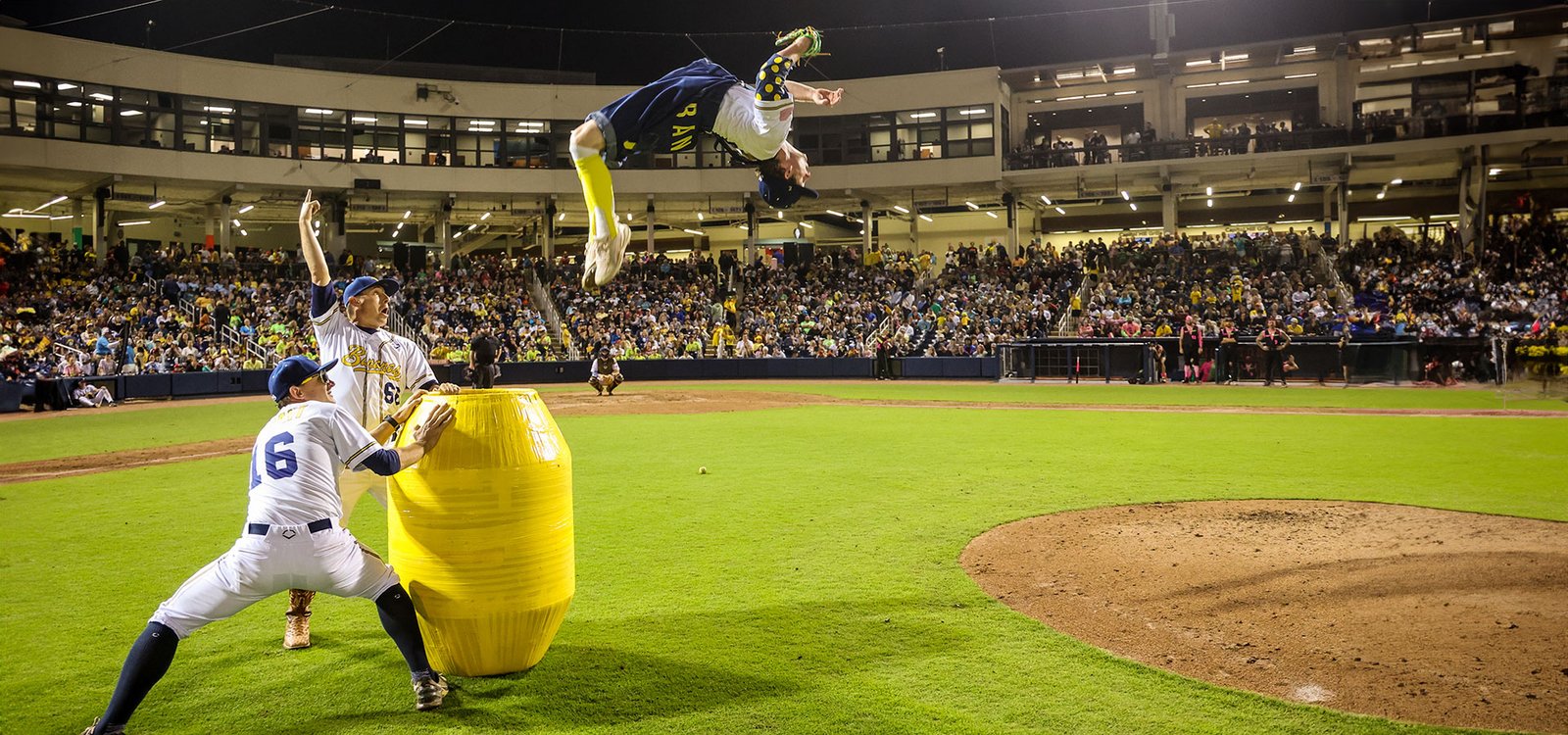 A Savannah Bananas player backflips over a giant yellow barrel in front of a packed stadium